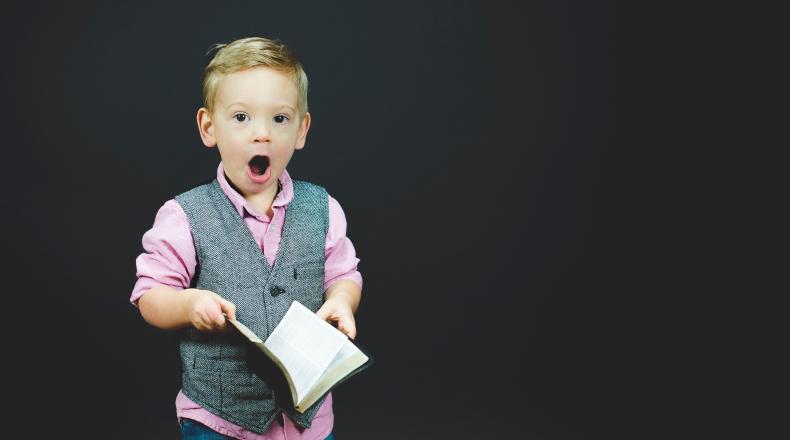 boy in pink shirt and grey vest reading a book and surprised 