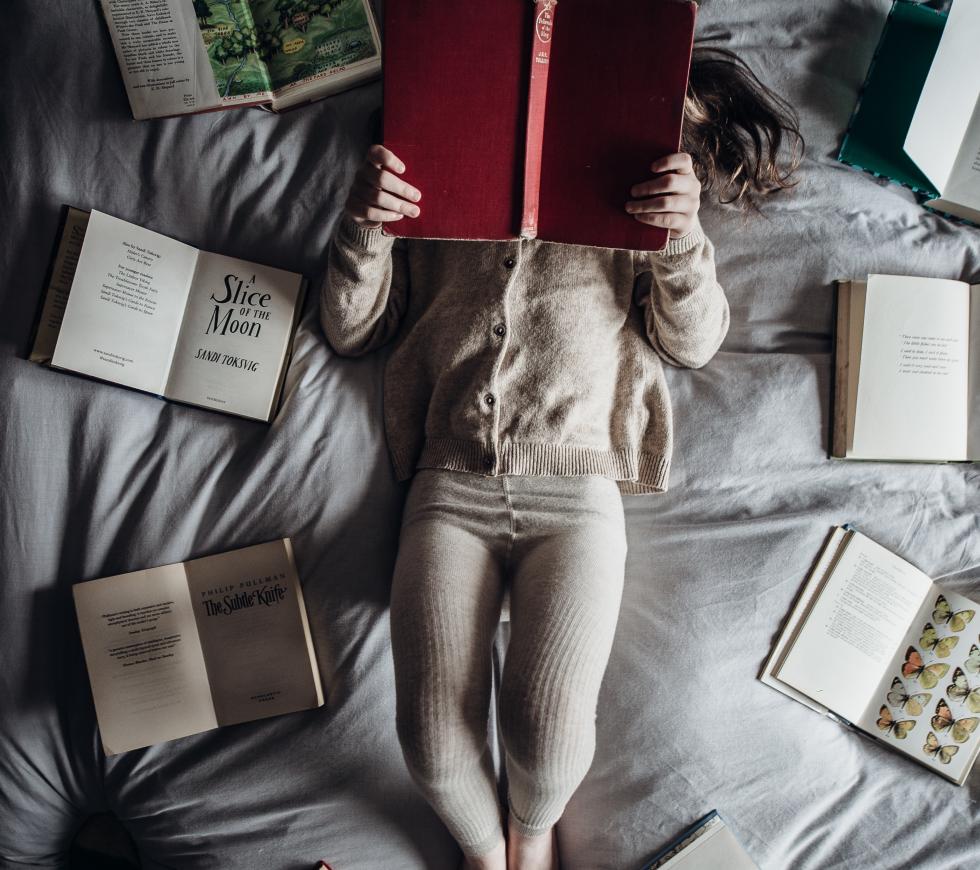A girl laying down reading a book while surrounded by other books. 